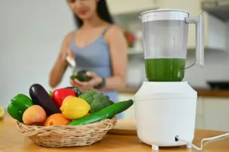 Blender and fresh vegetables on kitchen countertop with young woman cooking in background.