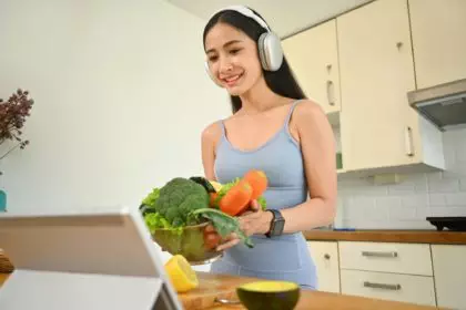 Happy young sports woman in headphone preparing food, making healthy salad in kitchen